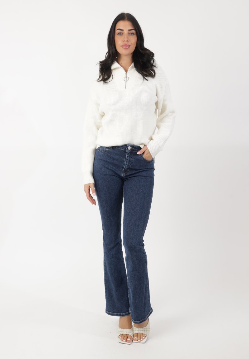 Pull en tricot blanc côtelé avec fermeture éclair, associé à un jean en denim bleu évasé. Le mannequin porte des sandales blanches à motifs. Fond uni.
