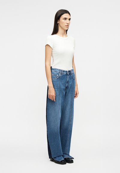 Γυναίκα όρθια, φοράει λευκό τοπ με κοντά μανίκια, μπλε τζιν παντελόνι με φαρδιά μπατζάκια και σκούρες πλευρικές λεπτομέρειες, και μαύρα παπούτσια, σε απλό ανοιχτόχρωμο φόντο.