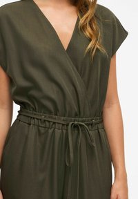 Mujer con un vestido envolvente sin mangas en color verde oliva, con un cinturón delgado atado a la cintura, mostrando la parte superior del torso y el brazo.