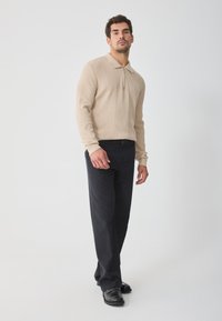 Beige gebreid vest met rits en ribbelstructuur, gecombineerd met navy broek en zwarte schoenen. Model poseert rechtop tegen een neutrale achtergrond.