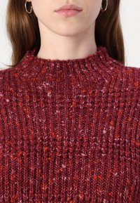 Πλεκτό πουλόβερ με ψηλό λαιμό, που διαθέτει μια υφασμάτινη σχέδιο σε αποχρώσεις βαθιού κόκκινου και πορτοκαλί. Διακριτική ανώμαλη υφή νήματος.