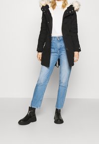 Svart parkajacka med pälsig huva, dragkedja och tryckknappar, kombinerad med ljusblå raka jeans och svarta ankelskor. Minimalistisk design.