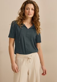 Kurzärmlige, dunkelblauen Bluse mit Kragen und einer Frontblende. Kombiniert mit beigen, Kordelzughosen mit Faltendesign.