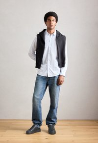 Junger Mann steht mit den Händen hinter dem Rücken, trägt ein weiß gestreiftes Hemd, einen schwarzen Pullover über den Schultern, blaue Jeans und schwarze Sneakers.