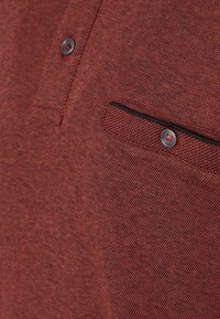 Punainen teksturoitu kangas, jossa on hienovarainen kuvio, varustettuna pienellä vetoketjullisella taskulla ja napin yksityiskohdalla. Lähikuva paljastaa ompelujäljen ja kudonnan.