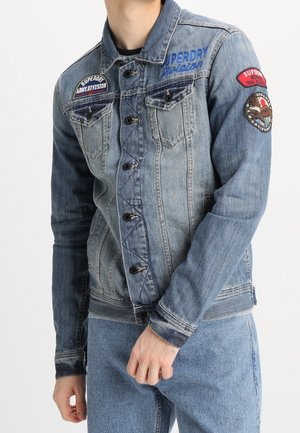 Mann trägt eine blaue Jeansjacke mit Knopfverschluss und bestickten Aufnähern auf Brust und oberem Ärmel, kombiniert mit blauen Jeans.