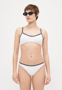 Hvit bikini med tynne stropper og svarte logo-detaljer på toppen og bunnen. Slett tekstur, minimalistisk design og kroppsnær fasong.