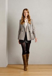 Blazer com um padrão zigzag multicolorido, blusa branca com um laço, saia mini preta, meias opacas pretas e botas castanhas até ao joelho.