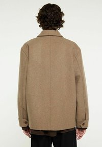 Brun ullrock med en lös passform, sned krage, stora ärmar och knäppta manschetter; slät textur och minimalistisk design.