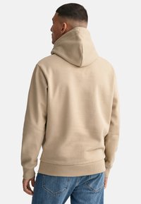 Felpa beige con un ampio cappuccio, polsini e orlo a coste. Tessuto morbido, vestibilità rilassata e design semplice. Indossata con jeans blu.