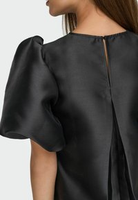 Mujer vistiendo una blusa de satén negra con mangas cortas abullonadas y un escote en la espalda con cierre de gancho.