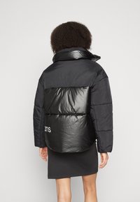 Svart puffjacka med en glänsande svart panel på baksidan, med hög krage och vit logodetalj vid nederkanten.