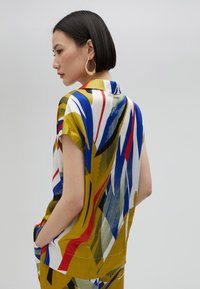 Krátky rukáv košele s odvážnym vzorom, obsahujúcim modré, červené, žlté a zelené abstraktné motívy na bielom pozadí. Hladká tkanina, detail goliera.