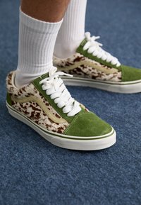 Personne portant des chaussettes blanches et des baskets à motifs verts et marron sur un sol en moquette bleue.