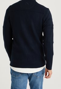 Marinblå stickad jacka med en skräddarsydd passform, långa ärmar och strukturerade sömmar. Lagd över en vit t-shirt, parat med ljusblå jeans.