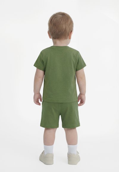 Παιδί που στέκεται με την πλάτη στραμμένη, φοράει πράσινο κοντομάνικο πουκάμισο, ταιριαστά σορτς, άσπρες κάλτσες και μπεζ παπούτσια, σε λευκό φόντο.