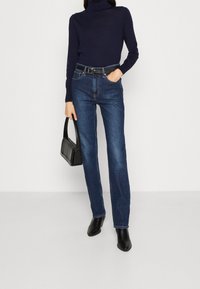 Marineblauwe coltrui, donkerblauwe jeans met rechte pijpen, zwarte riem en zwarte enkellaarsjes met hak, gecombineerd met een kleine zwarte handtas.