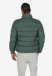 Chaqueta acolchada verde oscura con secciones acolchadas, cuello alto y logo en la parte posterior. El modelo lleva pantalones de color claro.