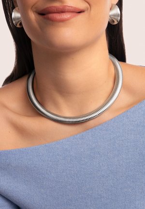 Un collier choker en métal argenté et des boucles d'oreilles créoles assorties présentent une texture striée et une forme cylindrique, portés avec un haut bleu à épaules dénudées.