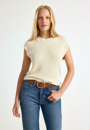Donna bionda con top crema senza maniche lavorato a maglia, jeans blu e cintura marrone, in piedi con una mano in tasca su sfondo bianco.