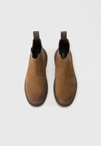 Barna suede Chelsea csizma sima felülettel, elasztikus oldalsó betétekkel, húzófülekkel és vastag gumitalppal. Standard bokamagasság.
