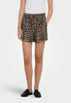 Kobieta ubrana w szorty w panterkę z wiązaniem w pasie, białą bezrękawnik oraz czarne wsuwane mokasyny, stojąca z jedną ręką w kieszeni.