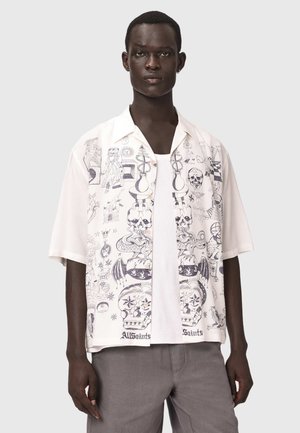 Junger Mann trägt ein weißes kurzärmeliges Hemd mit Knöpfen mit schwarzen detaillierten Schädel- und Schlangenillustrationen und graue Hose.