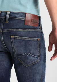 Jeans in denim blu scuro con una patch in pelle etichettata "MUSTANG" in vita, cuciture in contrasto e un design caratteristico della tasca posteriore.
