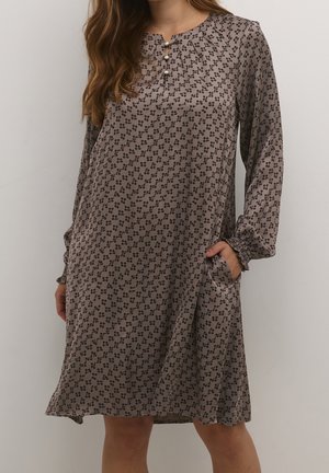 Femme portant une robe taupe à manches longues avec un motif à points noirs, des boutons en perle au niveau de l'encolure, et une main dans la poche latérale.