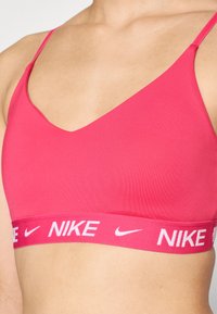 Rožs sports apakšveļa ar plānām siksnām, V veida dekoltē dizainu un platām elastīgām bandām ar baltu Nike logo tekstu. Gluda, stiepjama auduma tekstūra.