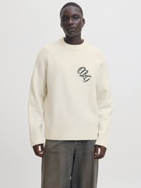 Cremfarbener Strickpullover mit rundem Halsausschnitt und gerippten Bündchen. Besitzt ein schwarzes gesticktes Logo auf der linken Brust. Getragen mit dunklen Jeans.