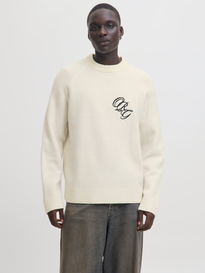 Cremfarbener Strickpullover mit rundem Halsausschnitt und gerippten Bündchen. Besitzt ein schwarzes gesticktes Logo auf der linken Brust. Getragen mit dunklen Jeans.