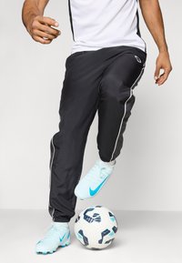 Schwarze Sporthosen mit weißen seitlichen Streifen, kombiniert mit hellblauen Fußballschuhen. Ein schwarz-weißer Fußball liegt auf dem Boden.