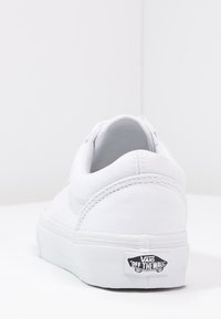 Bílé tenisky Vans z pohledu zezadu, ukazující šité detaily a logo "Vans Off The Wall" na podrážce na jednobarevném pozadí.