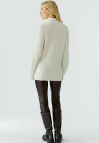 Pull overdimensionné de couleur crème avec un col haut, présentant une texture douce. Associé à un pantalon en cuir marron ajusté et à des bottes montantes.