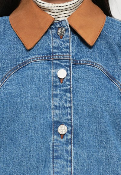 Τζάκετ από τζιν με καφέ φερμουάρ δέρματος. Διαθέτει μεταλλικά κουμπιά με εγχαραγμένο κείμενο. Μπλε ύφασμα με ορατές ραφές και απαλό υφή.