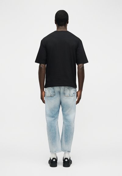 Άνδρας που φοράει απλό μαύρο κοντομάνικο μπλουζάκι, ανοιχτόχρωμο ξεβάμμενο τζιν, λευκές κάλτσες και μαύρα παπούτσια με λευκά λογότυπα φύλλου, στέκεται με την πλάτη γυρισμένη.