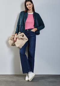 Námořnická saténová bomber bunda, růžové tričko s kulatým výstřihem, tmavé džíny s vysokým pasem a bílé tenisky. Drží dvě béžové fleecové tašky.