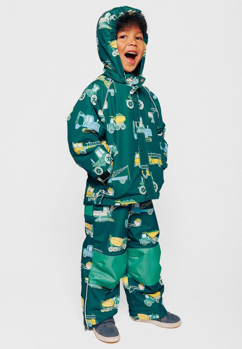 Giacca e pantaloni impermeabili verdi con motivo a veicoli da costruzione gialli e blu. Include un cappuccio e zip all'orlo dei pantaloni.