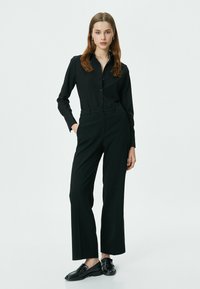 Zwarte button-up shirt met lange mouwen en een getailleerde pasvorm, gecombineerd met hoge taille, wijde zwarte pantalons en zwarte instapschoenen.