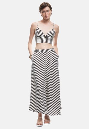 Nainen, jolla on ylhäältä mustavalkoinen raidallinen crop-top ja siihen sopiva pitkä hame, jossa on taskut, seisoo kädet taskuissa valkoista taustaa vasten.