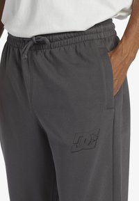 Gray drawstring sweatpants with an elastic waistband, side pockets, and a subtle logo print on the thigh. Soft fabric with a smooth texture.