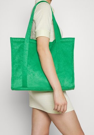 Personne portant une courte robe jaune à carreaux avec un grand sac fourre-tout vert sur l'épaule, sur un fond blanc uni.