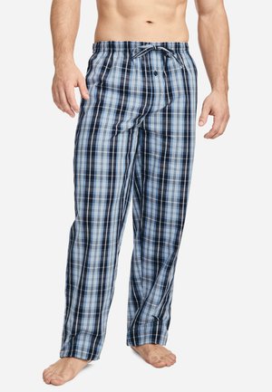 Mann trägt blaue und schwarze karierte Pyjamahosen mit Kordelzug in der Taille, steht barfuß vor einem schlichten weißen Hintergrund.
