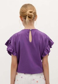 Lilla kortærmet skjorte med rynkede skulderdetaljer, der har en nøglehulsåbning bagpå, lavet af blødt stof.