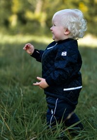 Black quilted bomber jacket with white accents, paired with black pants featuring white stripes. Child in a grassy field, side profile.