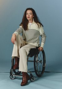 Jersey con rayas blancas y negras; tejido ligero y acanalado. Pantalones de pierna ancha beige y botas de tobillo de cuero marrón. Silla de ruedas, fondo gris.