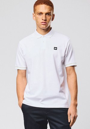 Joven con cabello rizado corto y rojo, vistiendo una camiseta tipo polo blanca con un pequeño logo negro y pantalones negros, de pie con las manos en los bolsillos.