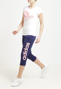 Valkoinen T-paita, jossa on monivärinen Adidas-logo; tummansiniset capripöksyt, joissa on suuri vaaleanpunainen "adidas"-printti; valkoiset urheilukengät.
