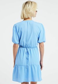 Vaaleansininen mekko, jossa on puffihihat, kapeat vyötäröt ja kerroksellinen hame. Valmistettu teksturoidusta kankaasta, jossa on sileä pinta. Säädettävä nauha takana.